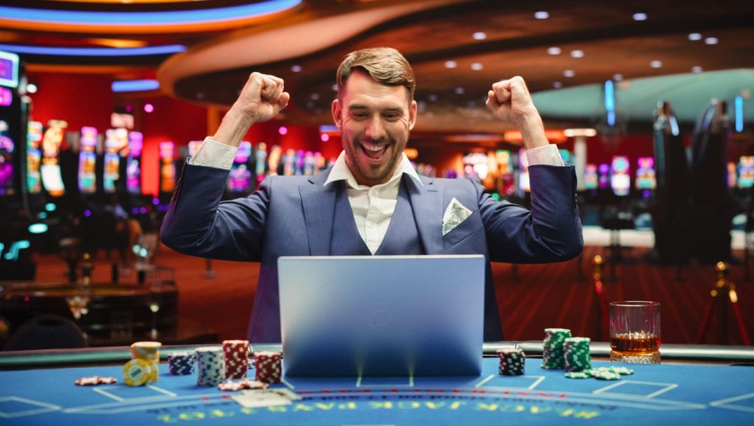 A person in a suit celebrates with both their hands in the air while sitting in front of a laptop and poker chip stacks at a casino table.