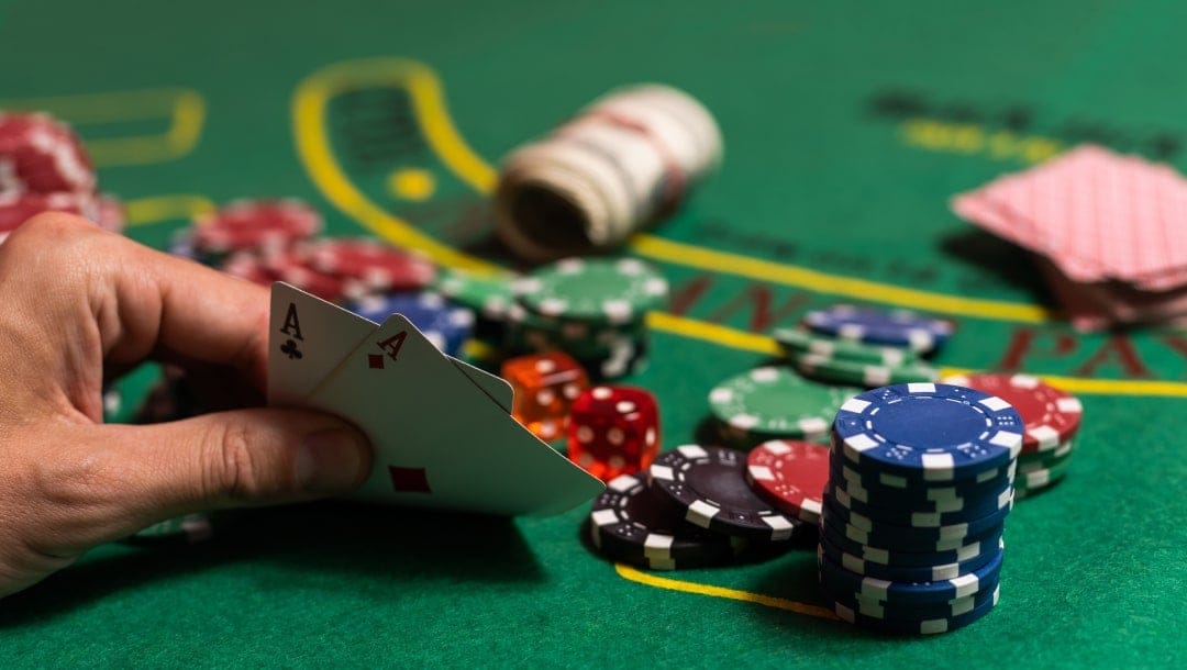 A closeup of a player using one hand to hold two Ace cards; Casino chips and a deck of cards are on the green blackjack table.