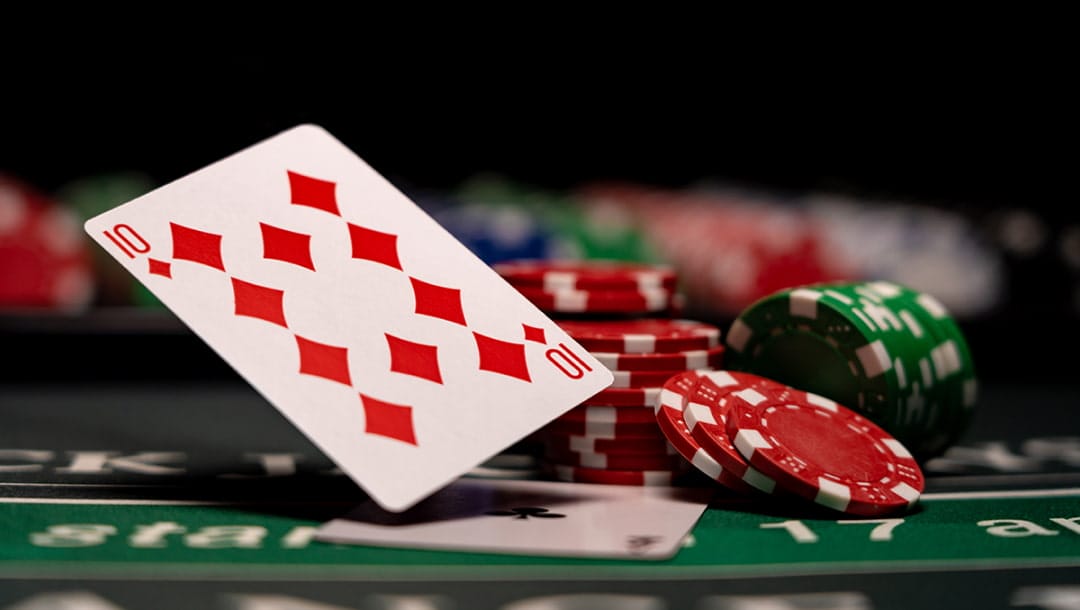A 10 of diamonds leans on top of multiple stacks of red casino chips. An ace of clubs is below it on a green casino table. Other stacks of chips are out of focus in the background.