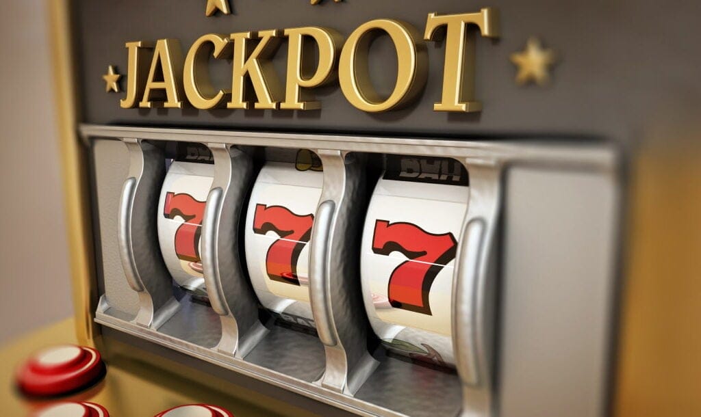 Close-up of a classic slot machine showing a winning combination of three red sevens on the reels, with the word 'JACKPOT' displayed prominently above in gold lettering. The machine has a sleek silver and gold design with red buttons on the front.