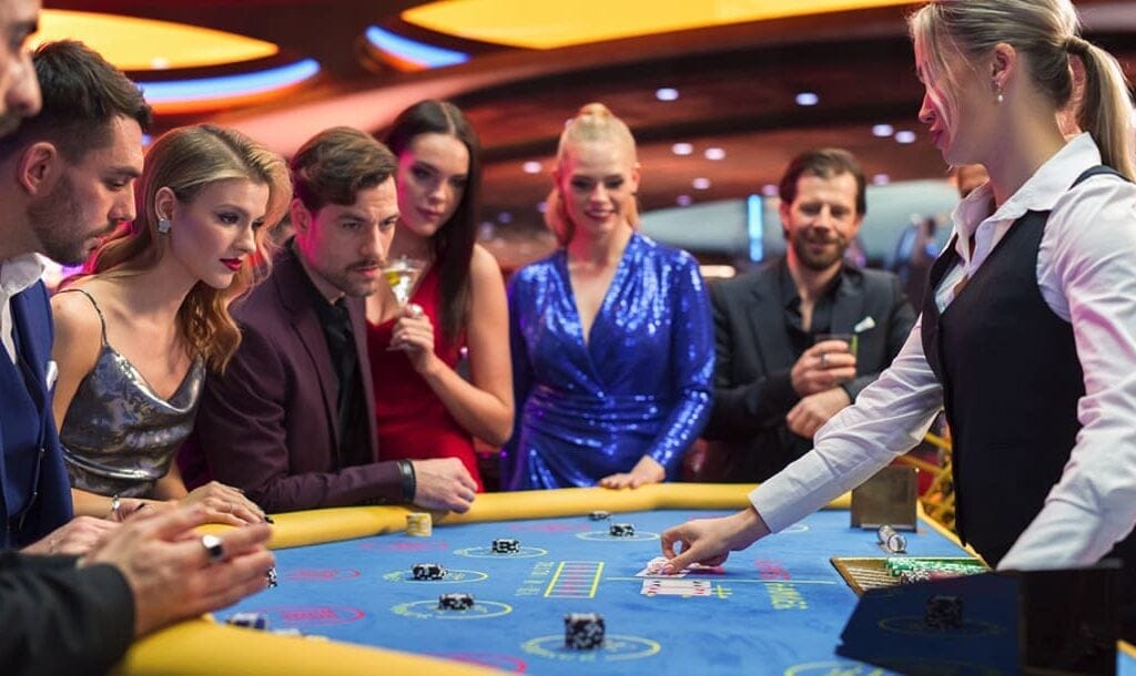 A group of people standing at a baccarat table. The baccarat dealer is touching playing cards on the blue baccarat table.