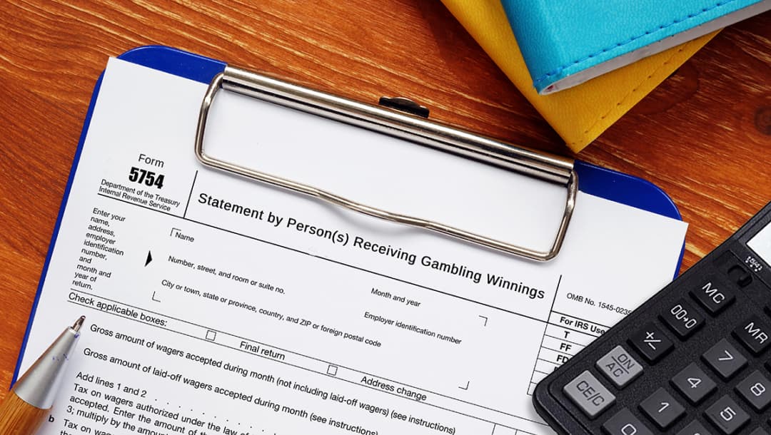 A gambling winnings form on a blue clipboard.The clipboard, calculator, pen and books are on a wooden surface.