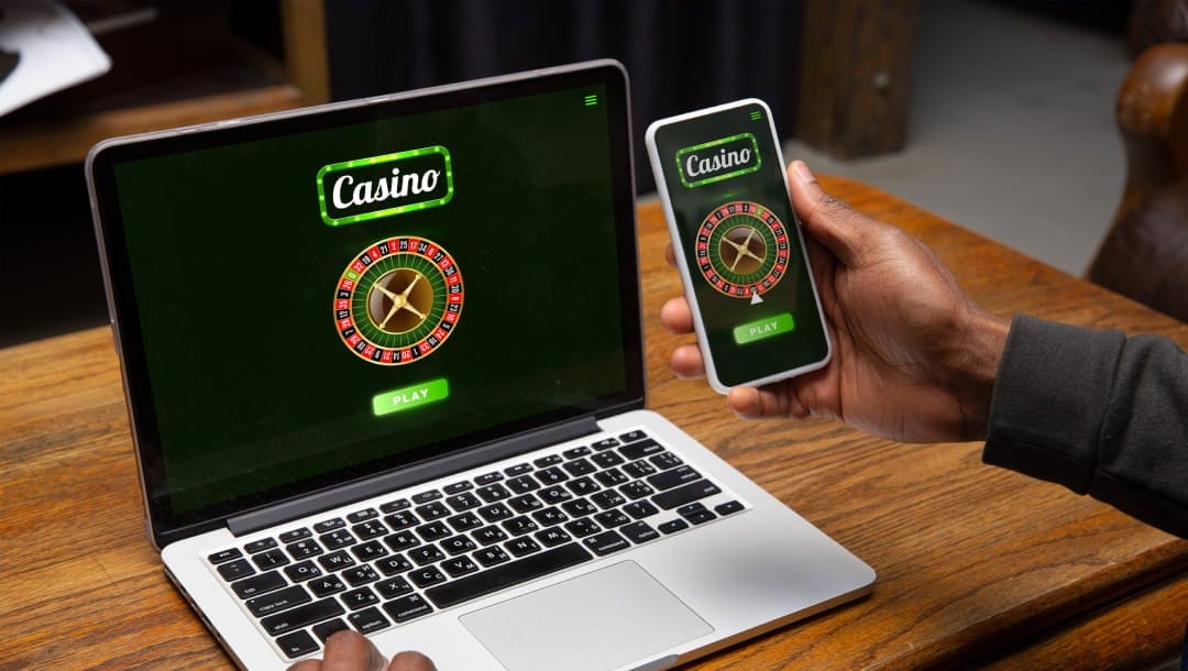 An over-the-shoulder view of a man holding a cellphone in his right hand while his left rests on the laptop in front of him. Both devices have online roulette loaded on the screen with a “play” button to start the games.