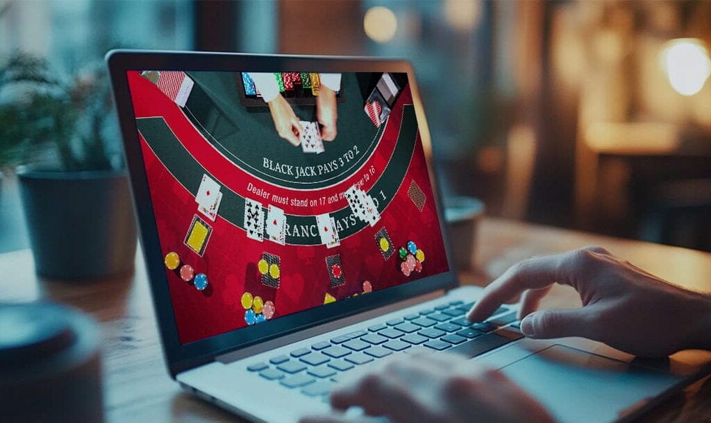 A photograph of a woman resting her hands on the keyboard of her laptop as she plays live dealer blackjack. The red blackjack table is visible on the screen from an aerial view, and the dealer is placing cards on the table.