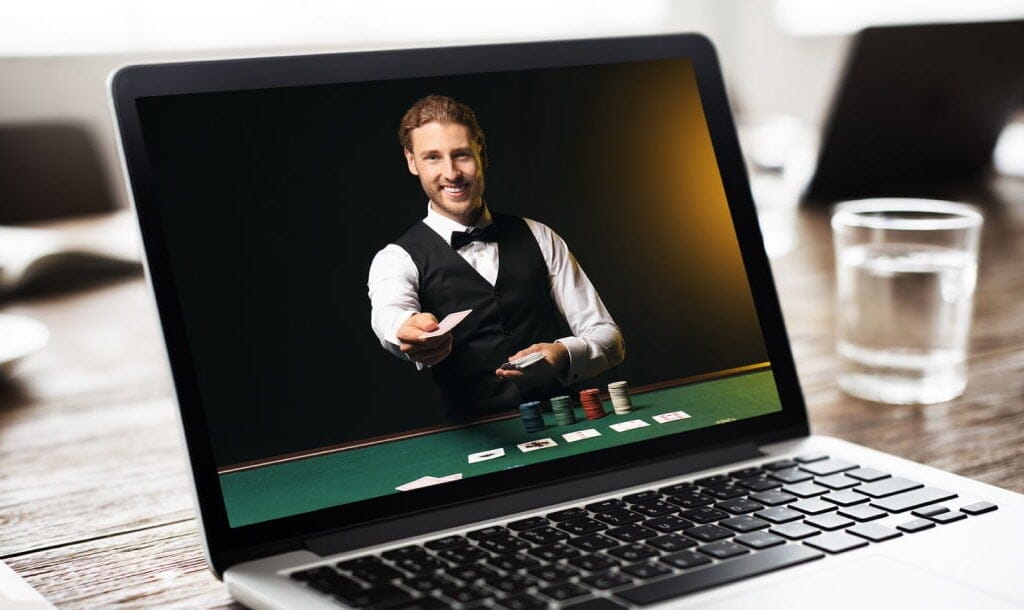 A photograph of a laptop, open, on a wooden table with a glass of water next to it. On the screen, there is a live dealer wearing a tuxedo, smiling while standing next to a poker table with stacks of chips and playing cards in front of him and cards in his hands.