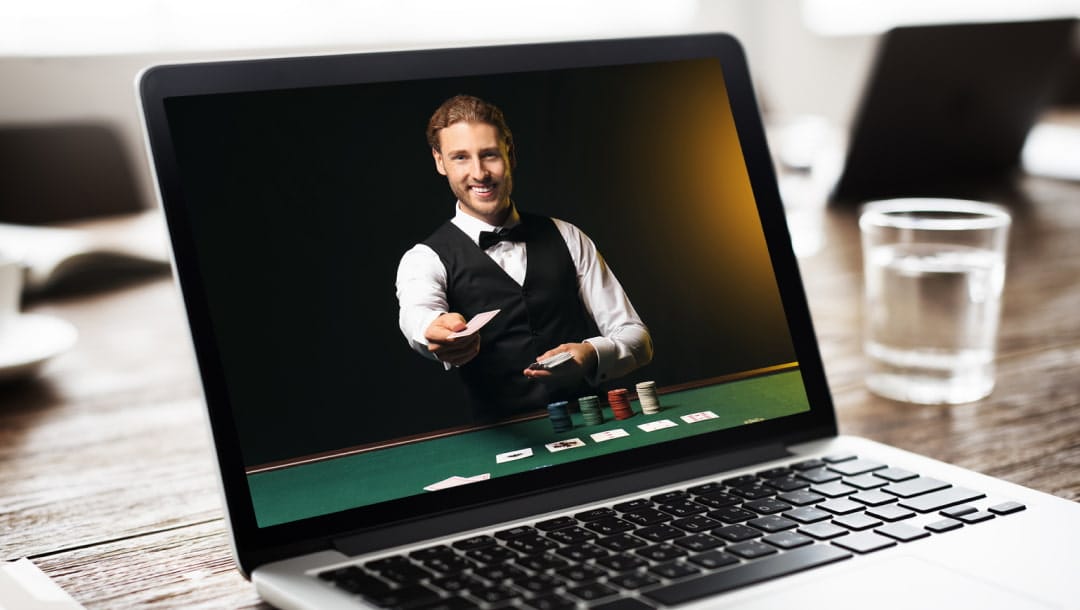 A photograph of a laptop, open, on a wooden table with a glass of water next to it. On the screen, there is a live dealer wearing a tuxedo, smiling while standing next to a poker table with stacks of chips and playing cards in front of him and cards in his hands.