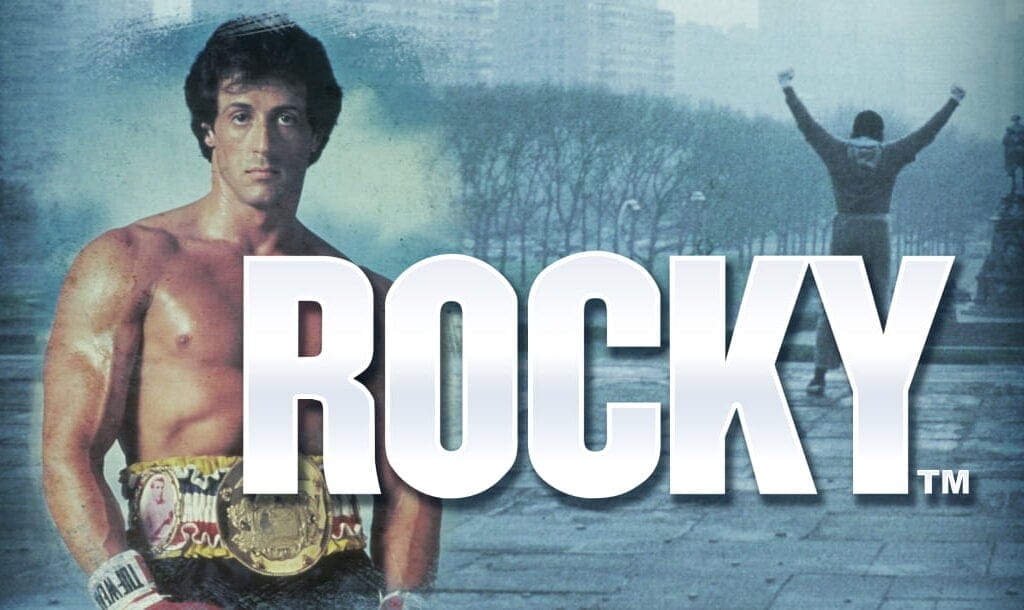 The Rocky title screen. The background is Rocky celebrating life at the top of the stairs of the Philadelphia Museum of Art. In the foreground is another image of Rocky with his title belt. The game title appears to the right of Rocky in a big, bold font.