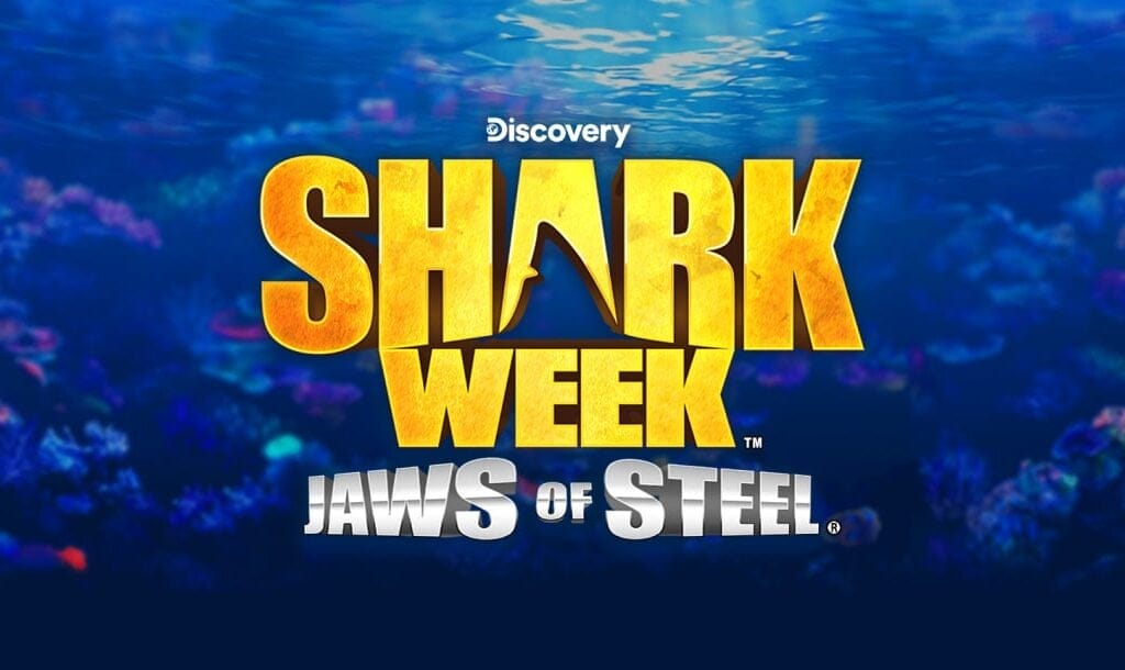 The title screen for Shark Week Jaws of Steel. The game title is spelled out in gold and silver letters. The “A” in “Shark” has a cutout of a shark fin in it. The background is an underwater image where coral can vaguely be seen.