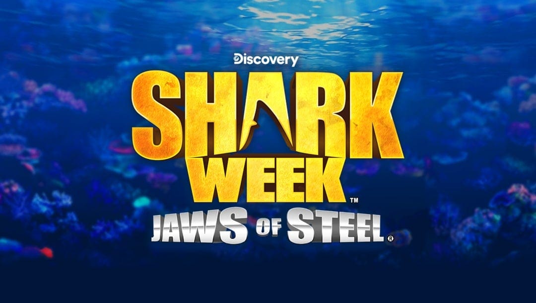 The title screen for Shark Week Jaws of Steel. The game title is spelled out in gold and silver letters. The “A” in “Shark” has a cutout of a shark fin in it. The background is an underwater image where coral can vaguely be seen.