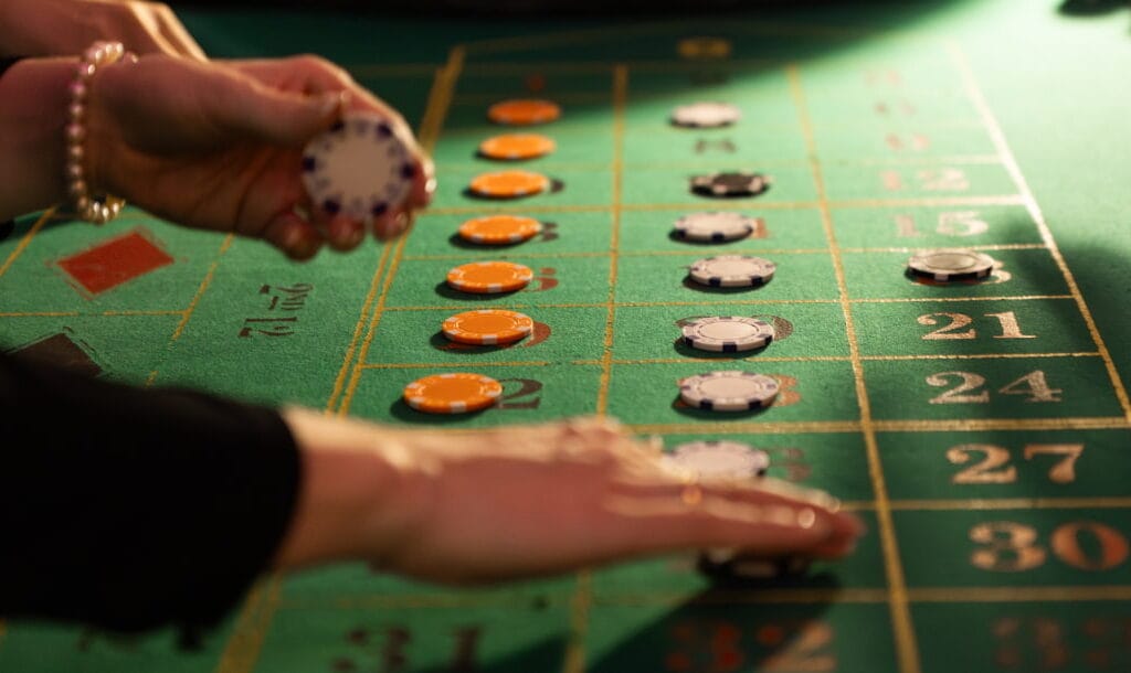 A close-up of a woman’s hands as she adjusts her bet at the roulette table. Her right hand is placing a chip on the table while she holds her remaining chips in her left hand. She is wearing a pearl bracelet and black shirt. Various bets are already on the roulette table.