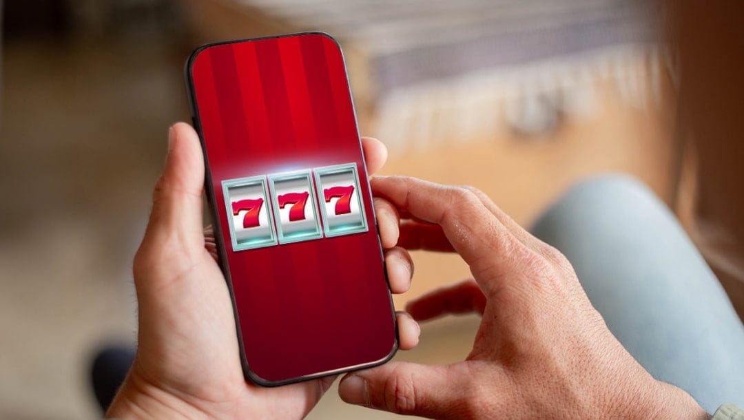 A person holding a smart phone with slot reels on it. There are three red 7s on the slot reels.