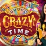 Promotional image for the live casino game "Crazy Time." A smiling game show host in a yellow and black circus-style jacket stands holding a gold microphone. Behind her is a large colorful spinning wheel with various numbers and bonus segments, along with vibrant elements like a blue train, gold coins, and a pink flamingo with a top hat, creating a lively, carnival-like atmosphere.