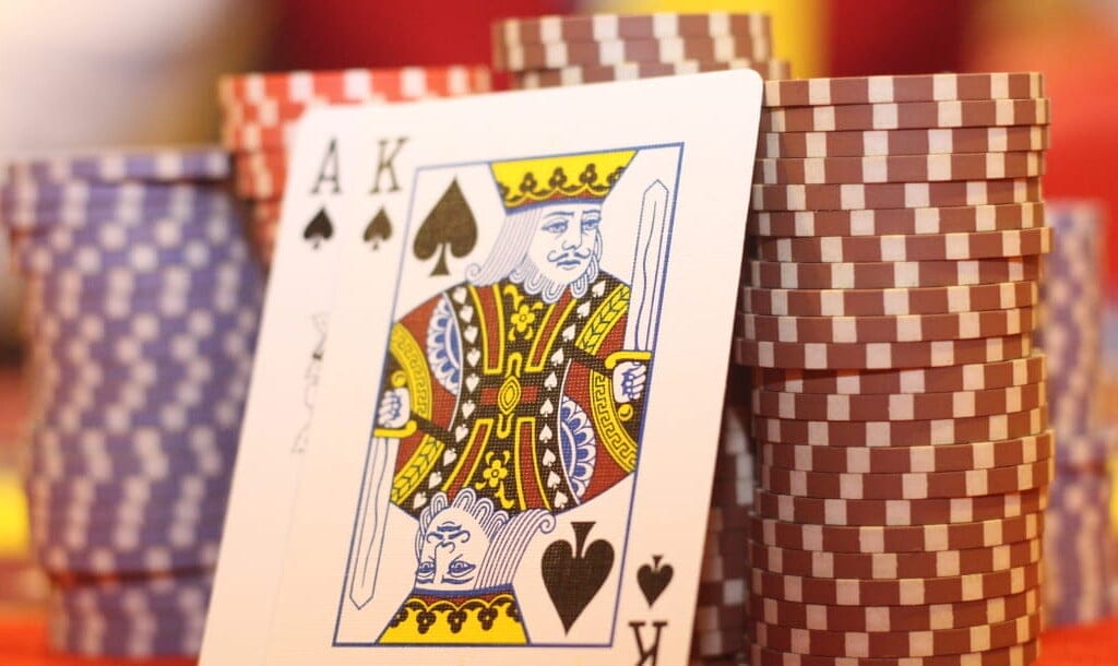 A close-up image of an Ace and King of Spades leaning against neatly stacked casino chips in shades of purple and brown. The cards and casino chips are set against a blurred casino background.