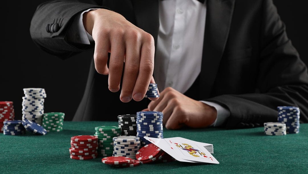 A person in a tuxedo seated at a green poker table stacking blue casino chips, with various colored chip stacks and two face-up cards visible in front.
