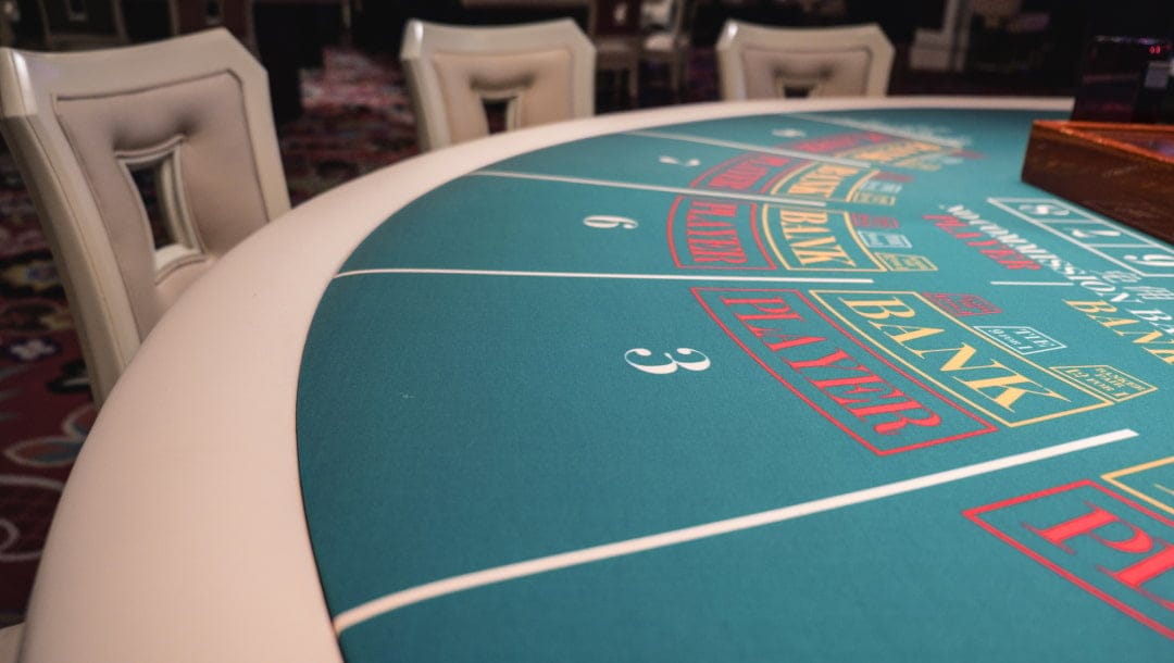 A close-up of a green baccarat table with white edging. There are white and grey chairs around the game table.