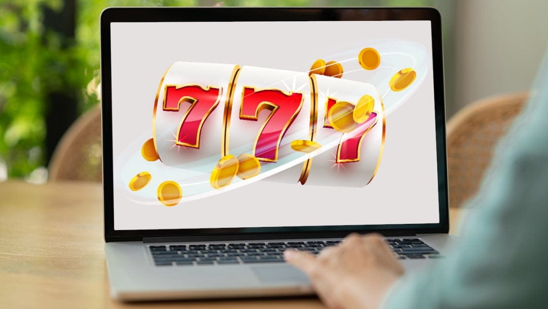 A person using a laptop, with the screen displaying a stylized slot machine reel featuring three large red sevens outlined in gold. The white and gold reels are surrounded by animated gold coins and swirling light trails. The laptop sits on a wooden desk with a soft-focus, green leafy background, suggesting a relaxed home environment for online gaming.